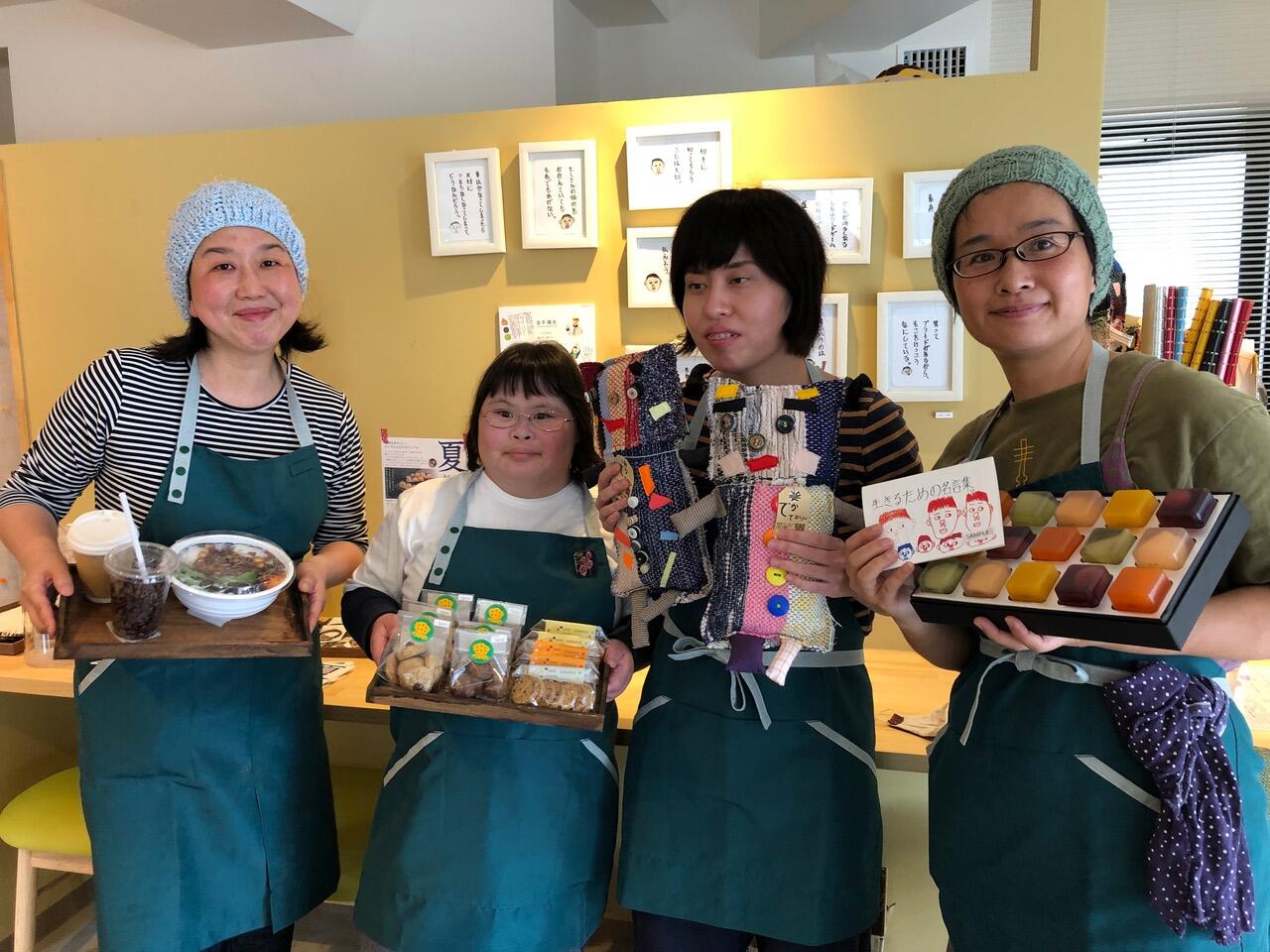 4人の店員がお菓子やご飯、グッズを持ちながらポーズ