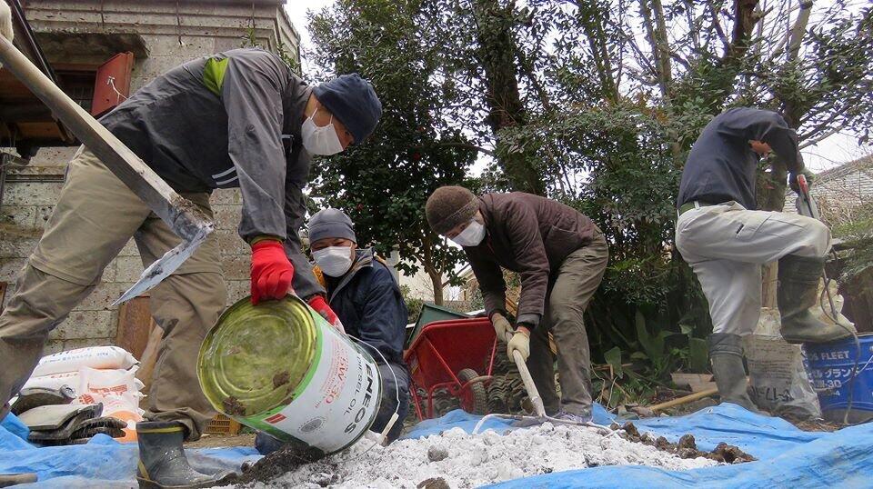 青のビニールシートの上に泥のような建築資材を広げて、それを作業員がスコップですくっている